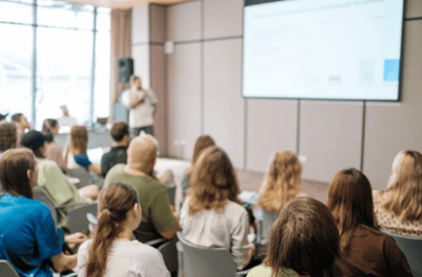 Publikum sitzt in einem hellen Konferenzraum und blickt auf einen Redner vor einer großen Präsentationsleinwand.